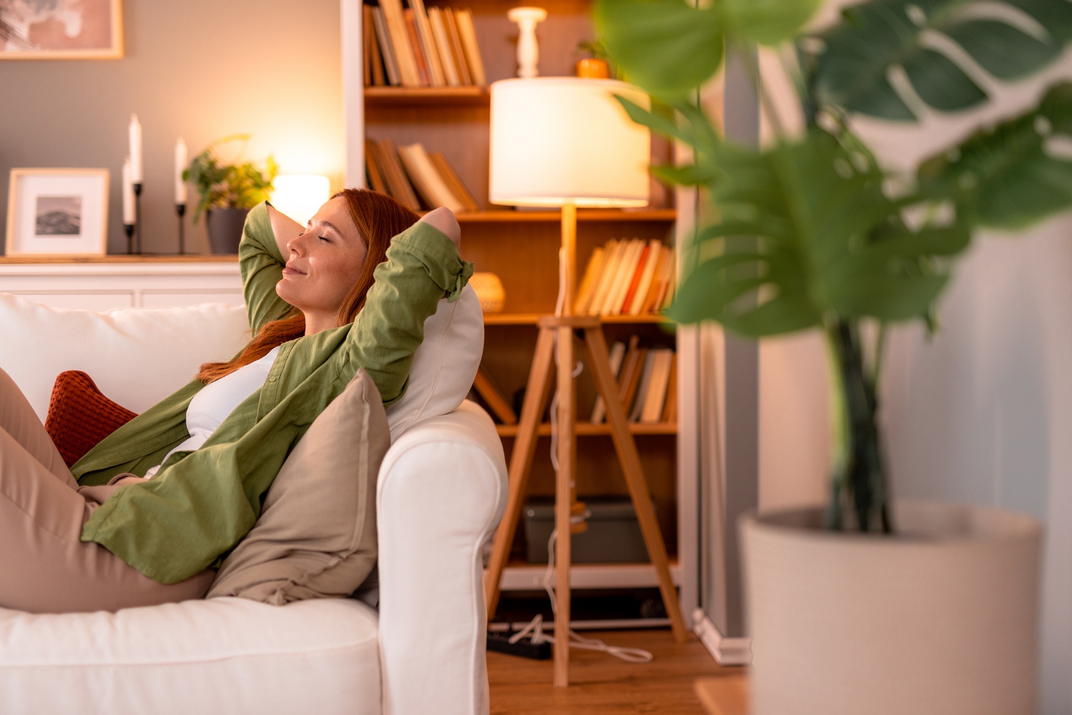 Femme rousse allongée sur un canapé blanc confortable, les mains derrière la tête, les yeux fermés, profitant d'un moment serein de détente dans un salon confortable et lumineux