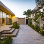 Allée pavée bordée de tropicale et de bâtiments en bois modernes, dans un quartier résidentiel ou entretenu bien. Décor végétal luxuriant avec palmiers et plantes fleuries, éclairé par douce lumière du soir, création ambiance chaleureuse accueillant