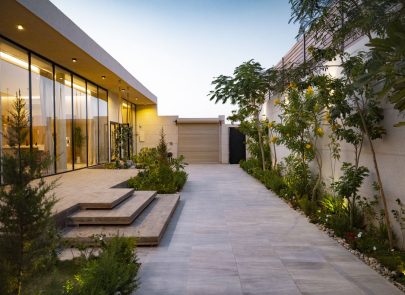 Allée pavée bordée de tropicale et de bâtiments en bois modernes, dans un quartier résidentiel ou entretenu bien. Décor végétal luxuriant avec palmiers et plantes fleuries, éclairé par douce lumière du soir, création ambiance chaleureuse accueillant