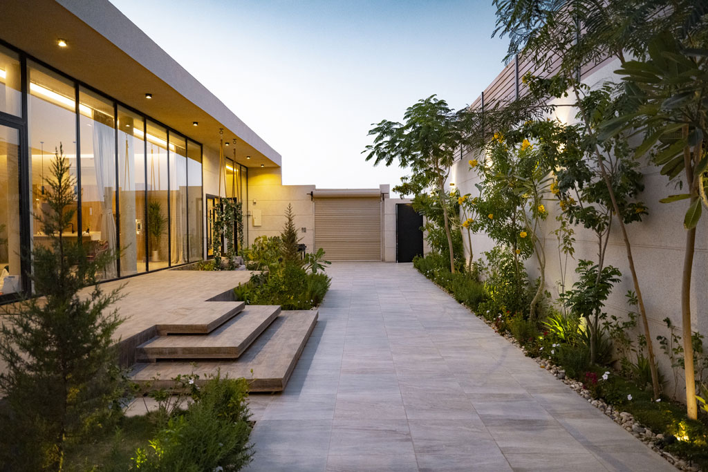 Allée pavée bordée de tropicale et de bâtiments en bois modernes, dans un quartier résidentiel ou entretenu bien. Décor végétal luxuriant avec palmiers et plantes fleuries, éclairé par douce lumière du soir, création ambiance chaleureuse accueillant